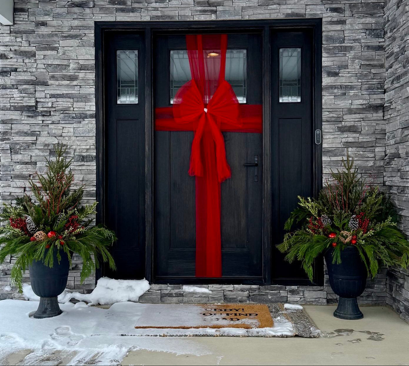 Black door with a large red bow, flanked by decorative plants on a stone porch.