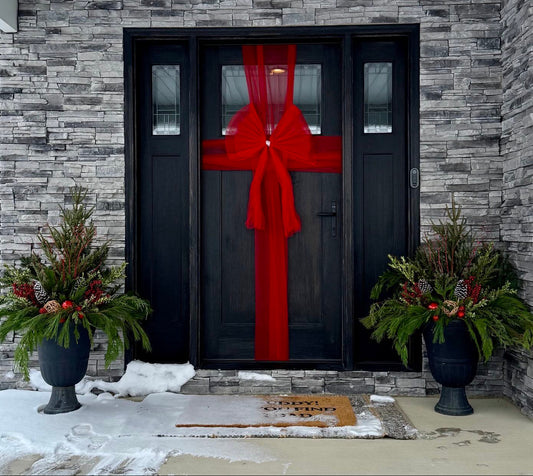 Black door with a large red bow, flanked by decorative plants on a stone porch.